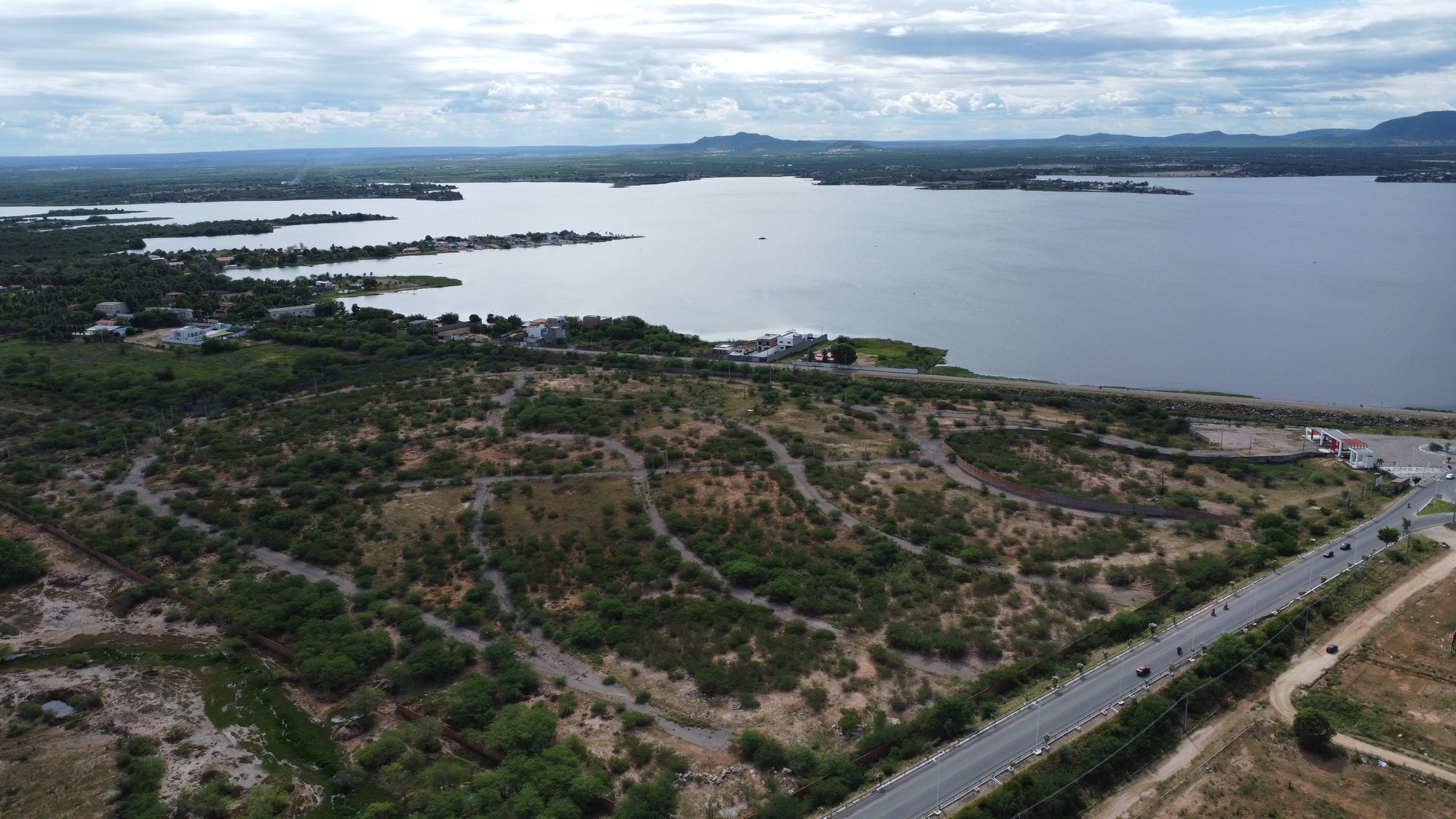 Vista aérea de uma área extensa às margens de um lago, com vegetação esparsa, traçados de vias no solo e poucas construções no entorno.